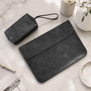 Op de afbeelding: Een donkergrijze leren laptoptas met een bijpassend klein etui. De laptoptas heeft een klepsluiting. Het etui heeft een ritssluiting en een zwarte polsband. De items liggen op een marmeren oppervlak.