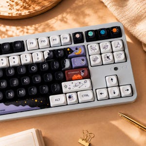 Può includere: Una tastiera meccanica con telaio grigio chiaro e un set di tasti personalizzati con grafiche a tema spaziale. I tasti neri, bianchi e viola presentano illustrazioni di astronauti, pianeti, stelle e razzi, oltre al testo ASTRONAUT.
