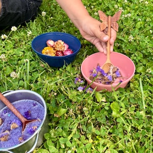 K&ouml;nnte beinhalten: Eine blaue Sch&uuml;ssel mit getrockneten Blumen, eine rosa Sch&uuml;ssel mit violetten getrockneten Blumen und ein silberner Metalltopf mit violetter Fl&uuml;ssigkeit und Blumen. Ein Holzl&ouml;ffel mit schmetterlingsf&ouml;rmigem Griff liegt in der rosa Sch&uuml;ssel.
