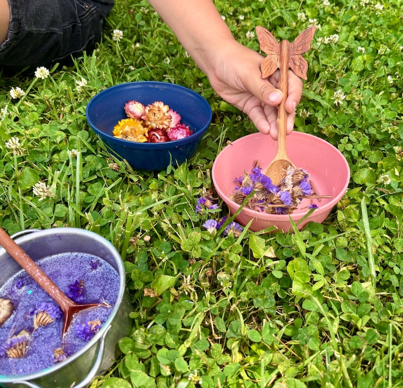K&ouml;nnte beinhalten: Eine blaue Sch&uuml;ssel mit getrockneten Blumen, eine rosa Sch&uuml;ssel mit violetten getrockneten Blumen und ein silberner Metalltopf mit violetter Fl&uuml;ssigkeit und Blumen. Ein Holzl&ouml;ffel mit schmetterlingsf&ouml;rmigem Griff liegt in der rosa Sch&uuml;ssel.