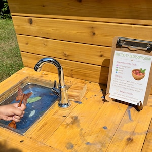 K&ouml;nnte beinhalten: Eine h&ouml;lzerne Outdoor-Spielk&uuml;che mit einem Edelstahlsp&uuml;lbecken, einem verchromten Wasserhahn und einem Klemmbrett mit einem Rezept namens Butterfly Blossom Soup. Das Rezept listet Zutaten wie Bl&uuml;tenbl&auml;tter, Kr&auml;uter, Gras und Wasser auf.
