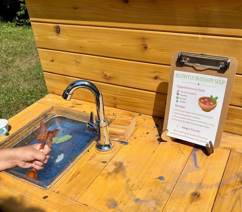 K&ouml;nnte beinhalten: Eine h&ouml;lzerne Outdoor-Spielk&uuml;che mit einem Edelstahlsp&uuml;lbecken, einem verchromten Wasserhahn und einem Klemmbrett mit einem Rezept namens Butterfly Blossom Soup. Das Rezept listet Zutaten wie Bl&uuml;tenbl&auml;tter, Kr&auml;uter, Gras und Wasser auf.