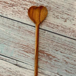 K&ouml;nnte beinhalten: Ein Holzl&ouml;ffel mit einer herzf&ouml;rmigen Laffe. Der L&ouml;ffel hat eine hellbraune Farbe mit einer sichtbaren, nat&uuml;rlichen Holzmaserung, die vertikal entlang des langen, schlanken Stiels und der herzf&ouml;rmigen L&ouml;ffelschale verl&auml;uft.