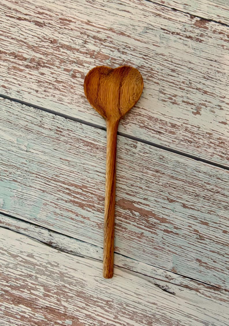 K&ouml;nnte beinhalten: Ein Holzl&ouml;ffel mit einer herzf&ouml;rmigen Laffe. Der L&ouml;ffel hat eine hellbraune Farbe mit einer sichtbaren, nat&uuml;rlichen Holzmaserung, die vertikal entlang des langen, schlanken Stiels und der herzf&ouml;rmigen L&ouml;ffelschale verl&auml;uft.