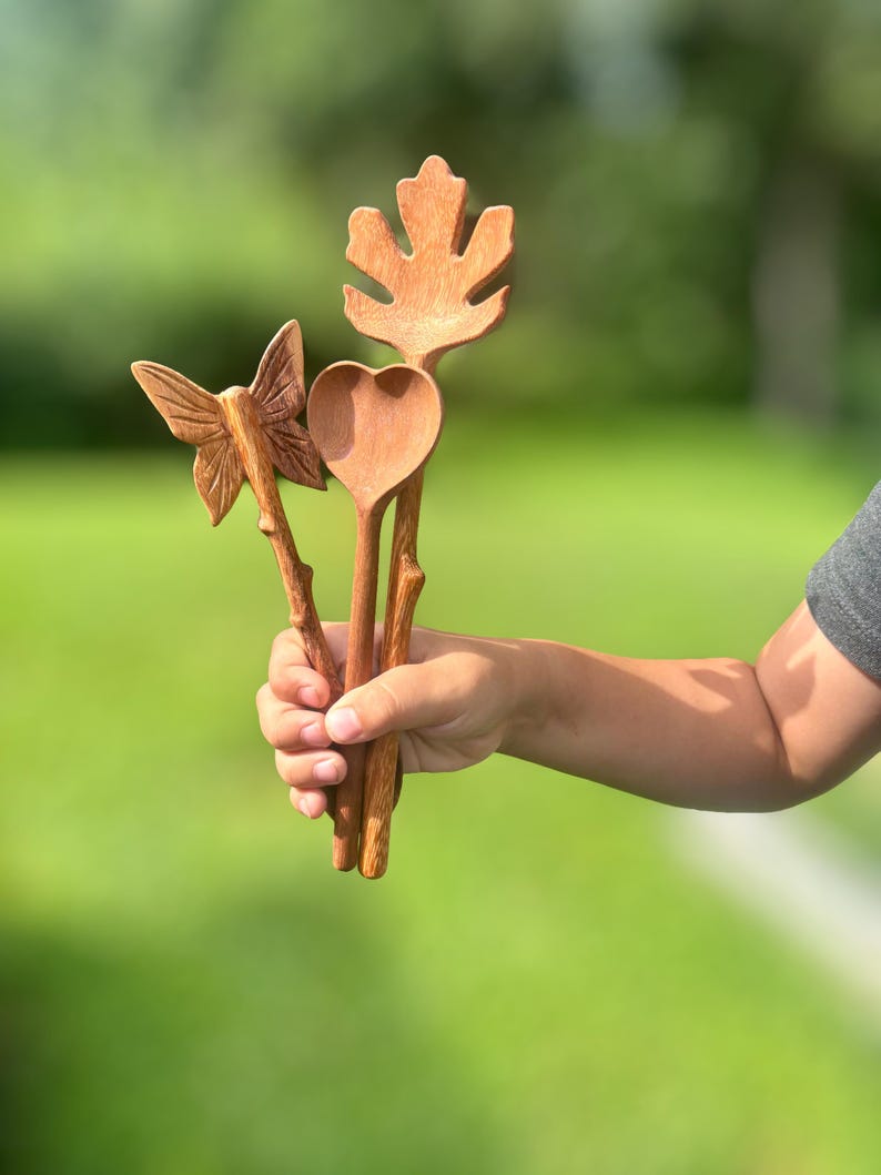 K&ouml;nnte beinhalten: Drei handgeschnitzte Holzutensilien in nat&uuml;rlichen Braunt&ouml;nen. Ein Utensil hat einen schmetterlingsf&ouml;rmigen Griff, ein anderes eine herzf&ouml;rmige Schale und das dritte ist mit einem gro&szlig;en Ahornblatt-Design versehen. Jedes St&uuml;ck hat einen strukturierten Griff.