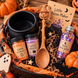 Op de afbeelding: Een Halloween-toverdrankset met een zwarte plastic ketel, een houten lepel met een uitgesneden pompoenhandvat, drie glazen potjes met labels en een kaart met de tekst Little Potion Maker, verpakt in een mandje met decoratieve elementen.
