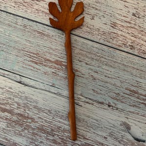 K&ouml;nnte beinhalten: Ein brauner Haarschmuck aus Holz mit einem geschnitzten Oberteil in Form eines f&uuml;nfzackigen Feigenblatts. Der Stiel des Haaraccessoires hat eine nat&uuml;rliche, leicht unebene Textur, die an einen kleinen Zweig erinnert, und eine glatte Oberfl&auml;che.