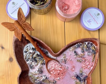 Könnte beinhalten: Eine schmetterlingsförmige Holzschale mit einer rosa Flüssigkeit, Blasen und getrockneten Blumen, dazu ein passender schmetterlingsförmiger Holzlöffel. Zwei durchsichtige Gläser sind vorhanden: eines mit getrockneten Schmetterlingsblüten und eines mit rosa Pulver.