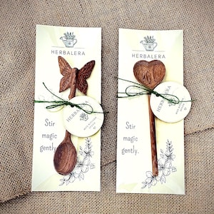 K&ouml;nnte beinhalten: Zwei Holzl&ouml;ffel auf wei&szlig;en Tr&auml;gerkarten mit dem Text HERBALERA und Stir magic gently. Ein L&ouml;ffel hat einen schmetterlingsf&ouml;rmigen Griff, der andere eine herzf&ouml;rmige Laffe. Beide sind mit gr&uuml;nem Garn und einem runden Etikett versehen.