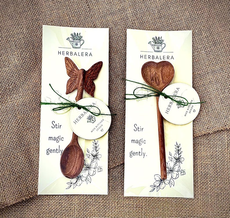 K&ouml;nnte beinhalten: Zwei Holzl&ouml;ffel auf wei&szlig;en Tr&auml;gerkarten mit dem Text HERBALERA und Stir magic gently. Ein L&ouml;ffel hat einen schmetterlingsf&ouml;rmigen Griff, der andere eine herzf&ouml;rmige Laffe. Beide sind mit gr&uuml;nem Garn und einem runden Etikett versehen.