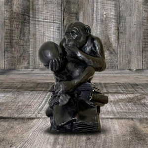 May include: A dark bronze-toned sculpture of a chimpanzee sitting on a stack of books. The chimpanzee is depicted in a contemplative pose, resting its chin on one hand while holding a skull in the other, with a textured finish throughout.