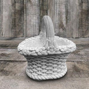 May include: A light gray concrete basket with a woven texture on the body and a decorative floral pattern along the top rim. The basket features a single arched handle extending from the center, with a rope-like design at the base.