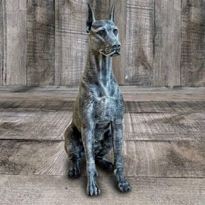 May include: A detailed statue of a Great Dane dog in a seated position. The sculpture is primarily gray with darker shading, giving it a textured appearance. The dog has a long neck, pointed ears, and a focused gaze. The statue is set against a wooden backdrop.