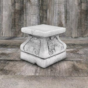 May include: A square stone pedestal with a flat top surface and a base featuring carved, textured scrollwork details on each side. The object is light gray in color with a weathered, matte finish and a solid, block-like structure throughout.