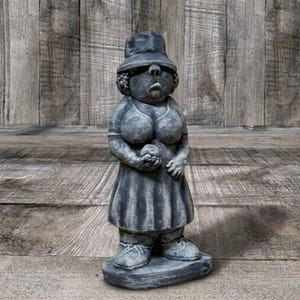 May include: A gray concrete garden statue of a woman wearing a hat and dress, holding a small object. The statue has a weathered appearance and stands on a rectangular base. The background is a wooden wall.