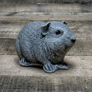 May include: A gray guinea pig figurine with detailed fur texture and brown eyes. The sculpture is posed in a seated position, with its paws visible. The figurine is placed on a weathered wooden surface, suggesting an outdoor or rustic theme.
