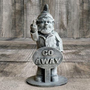 May include: A gray garden gnome statue with a beard and pointed hat, giving the middle finger. The gnome holds a sign that reads "GO AWAY". The statue is made of a stone-like material and stands on a circular base.