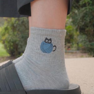 Könnte beinhalten: Ein Paar hellgraue Socken mit einem gestickten Motiv eines kleinen schwarzen Katers, der in einem blauen Wollknäuel sitzt. Die Socken haben eine gerippte Struktur und werden zusammen mit schwarzen Schlappen getragen.