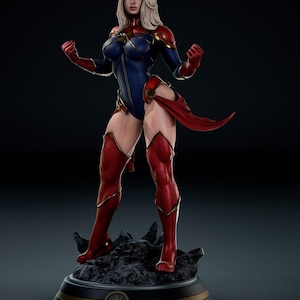 May include: A detailed figurine of a female superhero in a dynamic pose. She wears a blue and red bodysuit with gold accents, red boots, and a flowing red sash. Her blonde hair is styled, and she stands on a textured base with a star emblem.
