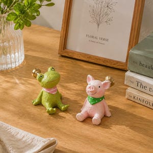 May include: Two decorative figurines: a green frog and a pink pig. The frog wears a pink scarf, and the pig wears a green scarf. Both have gold-colored accents and are sitting down. The figurines are on a wooden surface.