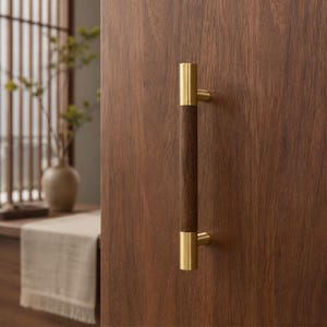 May include: A vertical door handle featuring a central dark brown wooden cylindrical grip flanked by two polished brass metal end caps. The handle is mounted onto a flat, smooth, dark brown wooden surface with a visible vertical wood grain pattern.