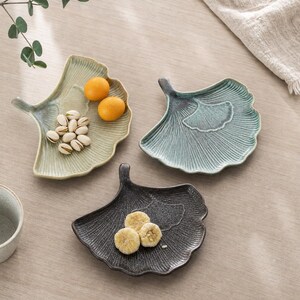 Puede incluir: Tres platos de cerámica con forma de hojas de ginkgo en verde claro, azul verdoso y gris oscuro. El plato verde claro contiene dos frutas pequeñas de color naranja y pistachos. El plato gris oscuro contiene tres rodajas de fruta seca.