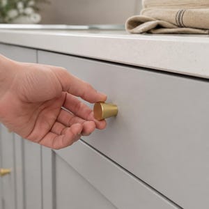 May include: A solid brass cabinet knob with a smooth, tapered cylindrical shape and a matte finish. The knob is mounted on a light grey drawer front, featuring a flat circular face and a slightly wider base that connects to the cabinet surface.