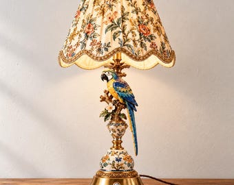 May include: A table lamp featuring a cream-colored pleated fabric shade with a floral and bird pattern. The base is a gold-toned metal stand with a sculpted blue, yellow, and white macaw parrot perched on a branch above a floral-patterned pedestal.