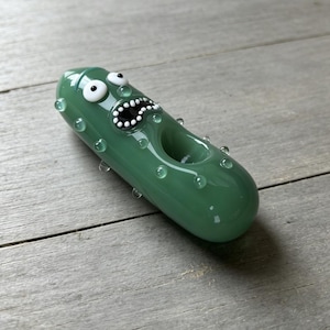 May include: A green glass pipe shaped like a pickle with a wide-eyed, open-mouthed face. The surface features small, raised glass bumps and a circular bowl opening on the side, crafted with smooth, translucent green glass and white accents.