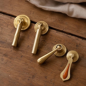 May include: Four brass furniture pulls with circular backplates. Three pulls feature cylindrical or tapered metal stems, while the fourth pull has a flat, teardrop-shaped stem with a smooth, glossy orange enamel inlay centered on the front surface.