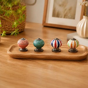 May include: Four decorative cabinet knobs with colourful, patterned ceramic spheres and bronze-toned bases are displayed on a wooden tray. The knobs feature designs in red, green, blue, and orange, adding a touch of whimsy to any furniture piece.