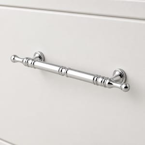 May include: A polished chrome drawer pull with a cylindrical bar design featuring decorative ribbed detailing near the ends and rounded finials. The hardware is mounted horizontally on a smooth, off-white surface with a subtle horizontal panel line.
