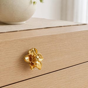 Könnte beinhalten: Ein goldfarbener Schubladenknopf aus Metall in Form einer blühenden Blume mit geschichteten Blütenblättern und einer strukturierten Mitte. Der Knopf ist an einer hellen Holzschubladenfront befestigt, die eine glatte Oberfläche aufweist.