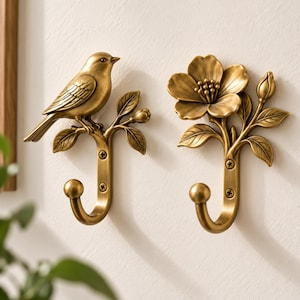 May include: Two antique brass-toned wall hooks. One hook features a bird perched on a branch with leaves and a bud. The second hook displays a large open flower with a bud and leaves. Both hooks have a curved base with two visible screw holes.