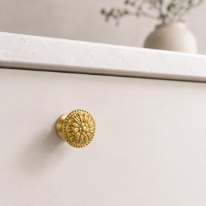 May include: A round, brushed gold cabinet knob featuring a raised, symmetrical floral petal design with a beaded outer edge. The knob is mounted on a smooth, light-coloured drawer front, showcasing a metallic finish and intricate detailing.