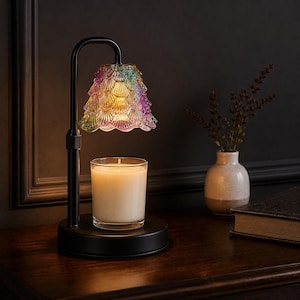 May include: A black metal candle warmer lamp featuring an adjustable arm and a scalloped, multi-colored glass shade with iridescent purple, green, and yellow hues. A clear glass jar candle with white wax sits on the circular base beneath the light.