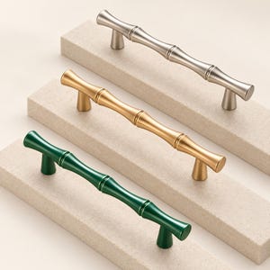 Puede incluir: Tres tiradores de armario con forma de tallos de bambú, cada uno con dos puntos de montaje. El conjunto incluye un tirador plateado, uno dorado y uno verde oscuro, todos con un acabado liso y uniforme y extremos redondeados.