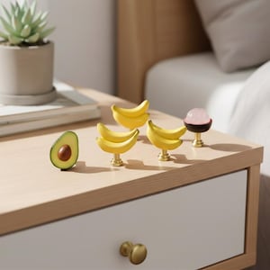 May include: A collection of decorative drawer pulls and a half avocado on a light wood surface. The drawer pulls include three sets of yellow bananas and a pink sphere on a black base. A small succulent plant in a pot is in the background.