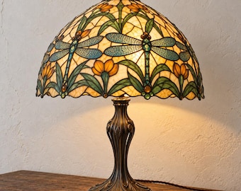 May include: A table lamp with a dome-shaped stained glass shade featuring a dragonfly motif in blue and green, accented by orange floral patterns. The lamp has a dark bronze-toned metal base with a curved, ornate stem and a circular foot.