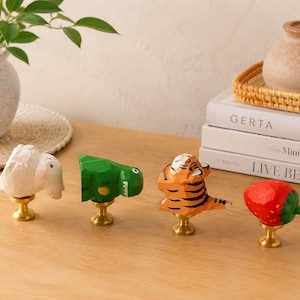 May include: Four decorative wooden knobs, each with a different animal or fruit design. The set includes a white rabbit, a green dinosaur, an orange tiger, and a red strawberry. Each knob is mounted on a gold-coloured base, ideal for furniture.