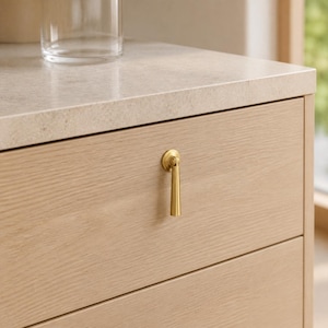 May include: A light-colored wooden drawer featuring a vertical, brushed brass pendant pull handle. The drawer is topped with a smooth, speckled off-white stone surface. The wood grain is visible, running horizontally across the drawer front.