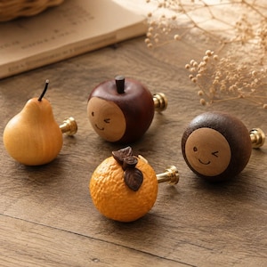 May include: Four wooden cabinet knobs in the shape of fruits. A pear, apple, orange, and a round brown knob with a smiling face. Each knob has a brass-colored base and is set against a wooden surface.