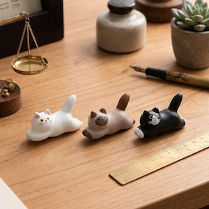 Puede incluir: Tres figuras de gato de cerámica en blanco, beige y negro, cada una en una pose juguetona. El gato blanco tiene orejas rosas y nariz amarilla, el gato beige tiene ojos azules y el gato negro tiene cara blanca. Una regla de latón está cerca.