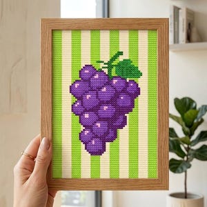 Peut inclure: Une œuvre d'art encadrée au point de croix représentant une grappe de raisins violets avec une feuille verte. Les raisins sont placés sur un fond à rayures vertes et crème, dans un cadre en bois brun clair. Une pièce décorative.