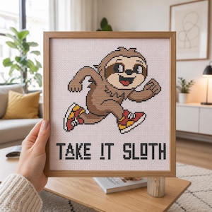 Peut inclure: Un motif de point de croix encadré représentant un paresseux de dessin animé marron qui court avec des baskets rouges et jaunes. Le paresseux a un large sourire et est placé au-dessus du texte noir TAKE IT SLOTH sur un fond à grille blanc.