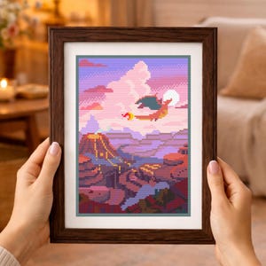 May include: A framed cross-stitch embroidery featuring a pixel art landscape with a flying orange dragon-like creature, a smoking volcano, and a large white moon set against a purple and pink sky with fluffy clouds, all encased in a dark wood frame.