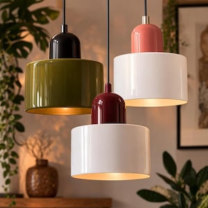 Peut inclure: Trois lampes suspendues modernes, de couleur vert olive, blanc et bordeaux, avec des dessus noirs, roses et bordeaux. Les abat-jours cylindriques ont une finition brillante et des intérieurs dorés, suspendus par des cordons noirs. Les lampes sont dans une pièce avec des plantes.