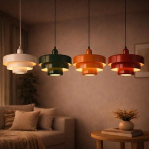 Peut inclure: Quatre lampes suspendues de couleurs blanche, verte, orange et rouge, chacune avec un design circulaire à plusieurs niveaux. Les lampes sont suspendues à des cordons noirs dans une pièce avec un canapé et une table d'appoint. Les lumières sont allumées, dégageant une lueur chaleureuse.