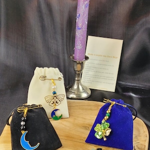May include: A lit purple candle in a silver holder, with three small velvet pouches and decorative pins. One pin features a butterfly, moon, and bead design. Another has a crescent moon. The third has a four-leaf clover.