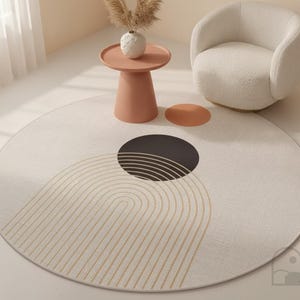 Puede incluir: Una alfombra circular con una base de color crema, que presenta un gran círculo negro y una serie de líneas finas y concéntricas de color marrón dorado que forman un arco. Un círculo más pequeño de color terracota se sitúa a la derecha del negro.